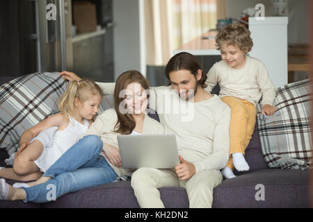 Happy family with kids using laptop sitting on sofa at home, smiling parents and son daughter relaxing on couch holding computer, man woman with child Stock Photo