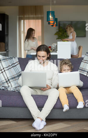 Dad and boy using laptops on sofa, kid imitating copying daddy holding computer sitting on couch while mother with daughter cooking spending time toge Stock Photo