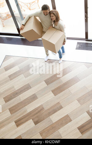 Happy couple coming into modern house through glass door holding boxes, smiling loving man and woman moving in new home with belongings, buying real e Stock Photo
