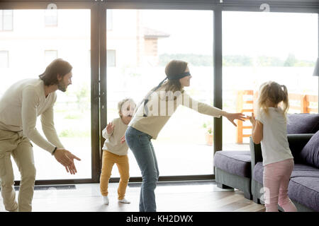 Blindfolded mother playing hide and seek game with father and kids hiding clapping hands at home, happy family having fun together on leisure in livin Stock Photo