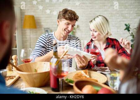 Portrait of young man and woman looking at digital tablet discussing photos while having dinner with friends sitting at big table with food Stock Photo