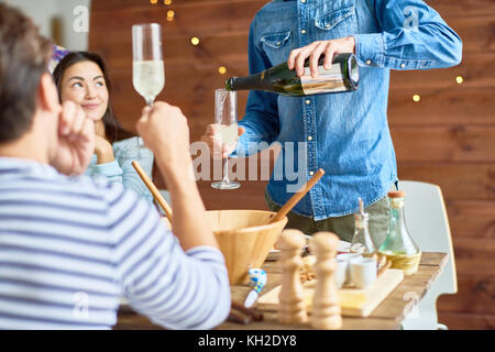 Group of happy young people wearing holiday caps celebrating at dinner table together, focus on man pouring champagne to glasses Stock Photo