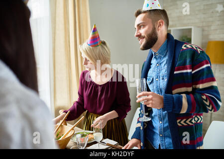 Group of happy young people wearing holiday caps celebrating Birthday with friends holding champagne glasses during party at home Stock Photo