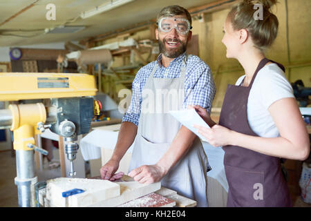 Portrait of bearded carpenter smiling happily talking to his female assistant in modern woodworking shop, standing next to drilling machine, copy spac Stock Photo