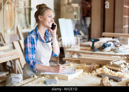 Portrait of modern young woman speaking by phone with client smiling and making notes in woodworking shop Stock Photo