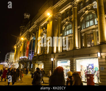 Manchester Royal Exchange Theatre building lit up at night  with shops and shoppers on St Annes Square, Manchester, UK