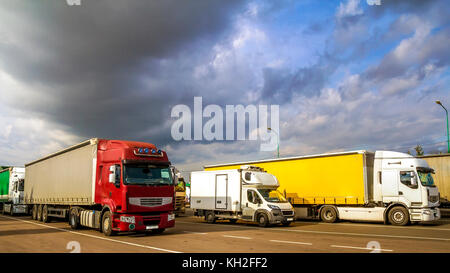 Colorful modern big semi-trucks and trailers of different makes and models stand in row on flat parking lot of truck stop in sunshine Stock Photo