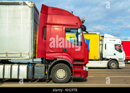 Colorful modern big semi-trucks and trailers of different makes and models stand in row on flat parking lot of truck stop in sunshine Stock Photo