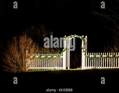 Arbor and gate pathway, white picket fence with Christmas garland at night, New Jersey, USA, Yule lights on vine arbor path archway garden arch Stock Photo