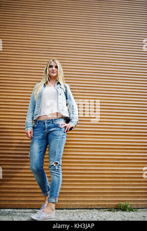 Blonde girl wear on jeans jacket with backpack posed against orange shutter. Stock Photo