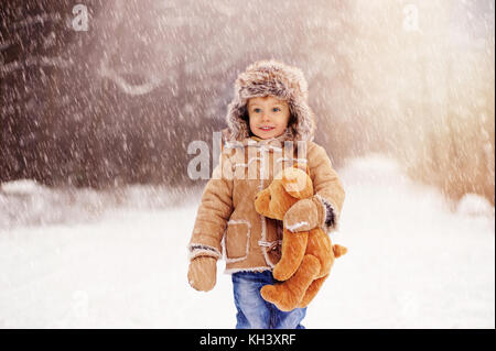 A little lovely smiling  boy wearing sheepskin cap standing on the snowy winter forest road and holding teddy bear. Outdoor waist up portrait. Stock Photo