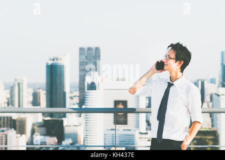 Young Asian businessman using mobile phone call on building rooftop, copy space on city view background. Business communication, concept Stock Photo