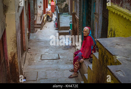 Varanasi city alleyway with old weathered buildings and an aged woman sitting at her doorway. Stock Photo