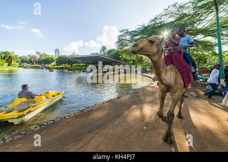 People on boats in boating lake and a camel ride with people on walk through Uhuru Park in late afternoon light, Nairobi, Kenya Stock Photo