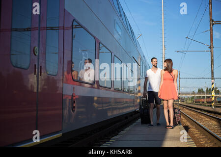 Happy young caucasian couple walking next to the train, pulling suitcases, wearing casual summer clothes Stock Photo