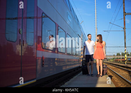 Happy young caucasian couple walking next to the train, pulling suitcases, wearing casual summer clothes Stock Photo