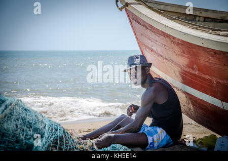 LOBITO, ANGOLA - MAY 09 2014: Unidentified Angolan fisherman sitting in front of red fishing boat at beach fixing nets Stock Photo