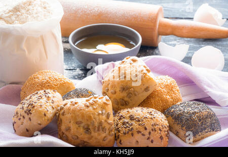 Food theme image with homemade bread rolls with sesame, poppy seeds, sunflower and flax seeds on a towel and the ingredients for their baking in the b Stock Photo