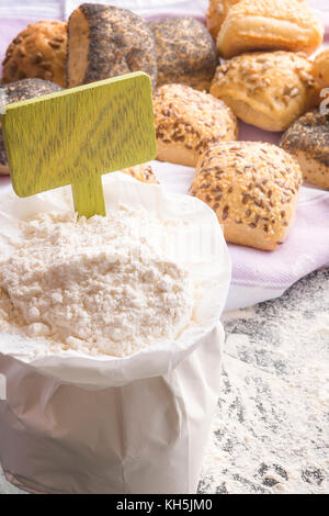 White paper bag full of flour with a wooden banner stick in it, homemade bread rolls decorated with seeds in the background on a kitchen towel. Stock Photo