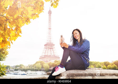 Happy young woman sitting on the embankment of Seine river with the Eiffel Tower on background and texting on smart phone Stock Photo