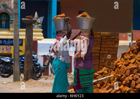 Two Indian women carry heavy bricks on their heads in traditional clothes. View from the back. Use of female labor at the building. Stock Photo