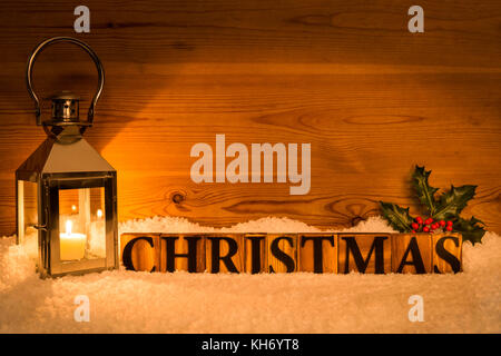 The word Christmas next to lantern with candle inside on a snow and wod background with holly Stock Photo