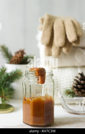 Caramel in a glass jar on a winter background with a rug or pullover, gloves, a cup, spruce branches and cones. Rustic style, selective focus. Stock Photo