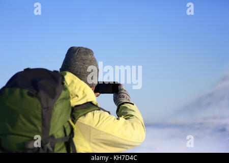 Back view of unrecognizable male in hat and cout with fur hood carrying stylish backpack taking picture of amazing winter landscape using copyspace mo Stock Photo