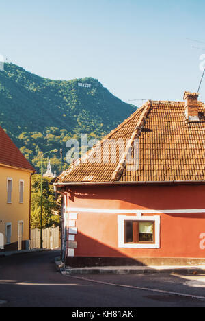A street with old colorful medieval vintage traditional romanian houses with tile roofs and a mountain with the sign of Brasov at the background in th Stock Photo