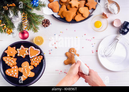 Woman hands holding a culinary bag with meringue cream inprocess of decorating gingerbread man cookies. White wooden table with ingrediens and decoration details. Christmas background concept. Stock Photo