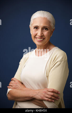 Photo of cheerful mature woman in beige tshirt, standing with crossed hands, looking at camera, over dark blue background Stock Photo