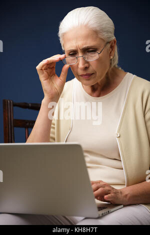 Image of serious old woman sitting over dark blue background using laptop computer. Looking aside. Stock Photo