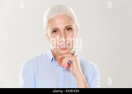 Close-up photo of elegant cherrful old lady, holding her chin, looking at camera, isolated over white background