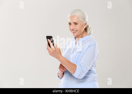 Mature concentrtated grey-haired woman using smartphone and smiling isolated over white Stock Photo