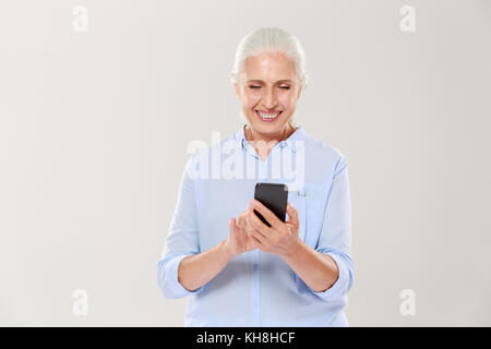 Mature concentrtated grey-haired woman using smartphone and smiling isolated over white Stock Photo