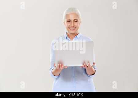 Cheerful mature grey-haired woman holding and using laptop computer isolated over white Stock Photo