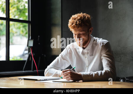 Photo of young smiling readhead bearded man, taking notes, while sitting at cafe Stock Photo