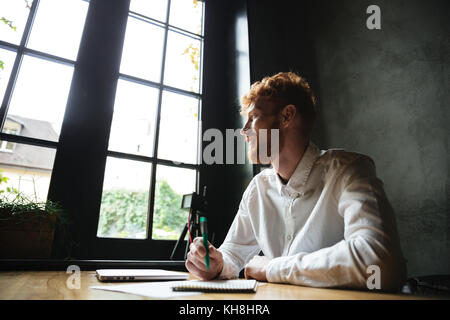 Photo of young smiling handsome readhead bearded man sitting on the workplace at home, looking at big window Stock Photo