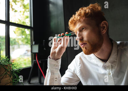 Close-up portrait of young concentrated readhead bearded man, holding green pen, looking at window Stock Photo