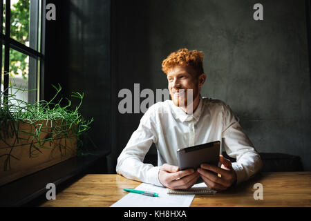Portrait of young cheerful ginger bearded man, sitting at wooden table, holding tablet, looking at window Stock Photo