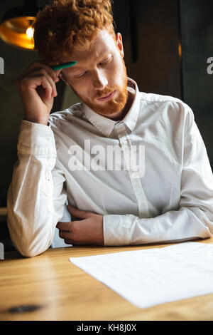 Portrait of concentrated readhead curly bearded man, reading papers , sitting at wooden table Stock Photo