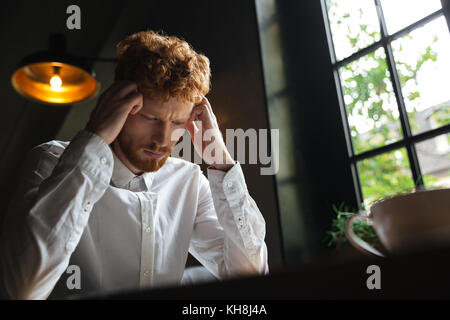 Close-up portrait of young readhead bearded overworked man in white shirt touching his head while sitting at office Stock Photo