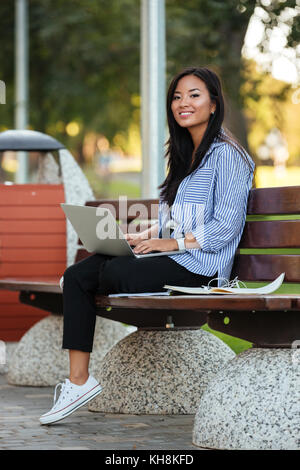Portrait of a happy pretty asian female student holding laptop computer while sitting on the bench at the park Stock Photo