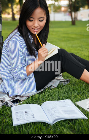 Close-up of asian female student, thinking with pencil in her mouth, sitting on grass in park Stock Photo