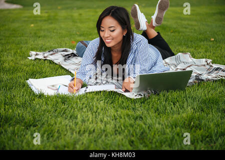 Beautiful asian female student, taking notes, lying on checkered plaid in park Stock Photo