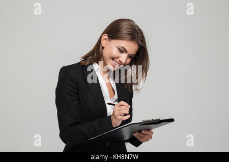 Portrait of a pretty satisfied businesswoman in suit signing documents while standing isolated over white background Stock Photo