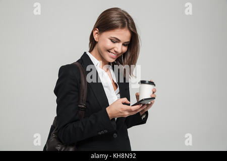 Portrait of a lovely smiling woman in suit using mobile phone while standing and holding coffee cup isolated over white background