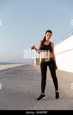 Full-length shot of young confident carefree lady in sportswear training near sea with jumping rope in morning Stock Photo