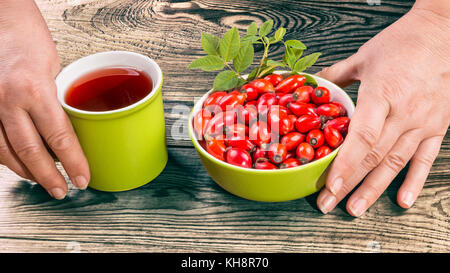Fresh fruits of wild rose and hip tea on brown wooden background. Hands holding bowl full of red rosehips and mug with medicinal fruit drink. Stock Photo
