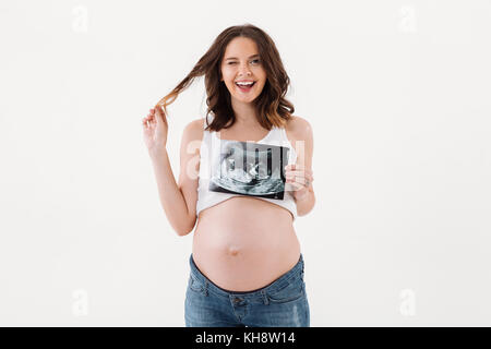 Photo of cheerful pregnant woman standing isolated over white background. Looking camera winking holding ultrasound scan of a baby. Stock Photo